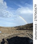 A vibrant double rainbow arching over a massive, raw open-pit mining excavation site. A striking contrast between the industrial, muddy earthwork and the beauty of natural atmospheric phenomena.