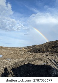 A vibrant double rainbow arching over a massive, raw open-pit mining excavation site. A striking contrast between the industrial, muddy earthwork and the beauty of natural atmospheric phenomena.