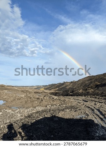 A vibrant double rainbow arching over a massive, raw open-pit mining excavation site. A striking contrast between the industrial, muddy earthwork and the beauty of natural atmospheric phenomena.