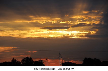 Vibrant and dramatic sunset featuring golden, orange, and dark clouds. Powerful sunbeams radiate from behind the cloud cover, creating a fiery sky effect. Minimalist silhouette of a suburban landscape and power lines at the bottom.