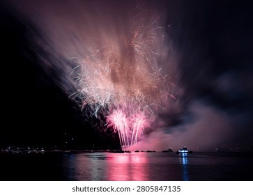 The vibrant fireworks explode over a calm water body, reflecting colorful lights in the night sky.