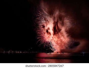 The vibrant fireworks explode over a calm water body, reflecting colorful lights in the night sky.