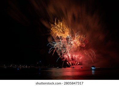 The vibrant fireworks explode over a calm water body, reflecting colorful lights in the night sky.