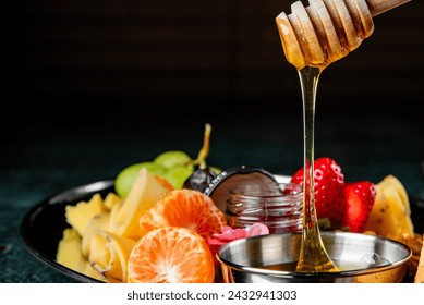 Vibrant fresh fruits and cheese on a plate, with golden honey drizzling from a wooden dipper, against a dark background - Powered by Shutterstock