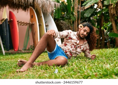 Vibrant gay man with flower in hair sits on grass, surfboards casual summer wear, relaxed. LGBT traveler enjoys tropical garden, represents diversity in leisure, confidence meet vacation bliss. - Powered by Shutterstock