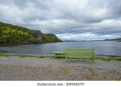 A Vibrant Green Bench And Picnic Table On A Beach. There's A Mountain Covered In Autumn Trees On One Side Of The Cove. The Beach Has Grass And Pebbles. The Water Is Calm And Smooth. The Sky Is Cloudy.