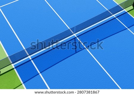 Vibrant high-angle view of an empty blue and lime green pickleball court with white boundary lines and a net. Bright sunlight casts sharp, dramatic shadows on the textured hardcourt surface.