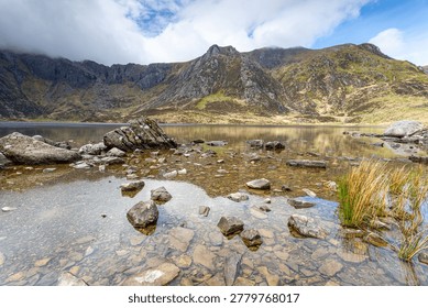 A vibrant, high-dynamic-range landscape featuring the crystal-clear waters of Llyn Idwal fringed by rich green summer vegetation. The stone-pitched hiking trail leads the eye toward the dramatic glacial cwm, with the rugged peaks of the Glyderau partially veiled in soft, white clouds.