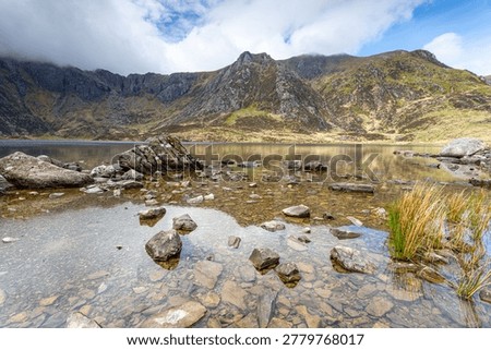 A vibrant, high-dynamic-range landscape featuring the crystal-clear waters of Llyn Idwal fringed by rich green summer vegetation. The stone-pitched hiking trail leads the eye toward the dramatic glacial cwm, with the rugged peaks of the Glyderau partially veiled in soft, white clouds.