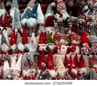 A vibrant holiday setup featuring rows of Christmas gnome figurines with long white beards and pointed hats in festive colors like red, gray, and white. Some are decorated with stars and hearts, while plush snowmen in scarves and winter hats add a playful touch. Nutcracker dolls and other seasonal characters peek from the background, creating a joyful and cozy atmosphere filled with handcrafted charm.