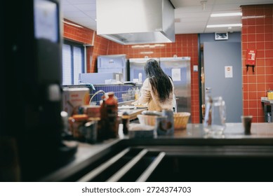In a vibrant kitchen setting within the Netherlands, a woman is focused on preparing food. The kitchen is bustling with activity, featuring red tiled walls and various cooking equipment in the background. Shelves are stocked with ingredients and utensils, while a variety of condiments are visible on the countertop. The atmosphere reflects a blend of professionalism and homey comfort, suggesting a space where culinary creativity flourishes.