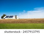 A vibrant landscape featuring a farm with a large solar panel array and several wind turbines against a clear blue sky, set in a plowed field.