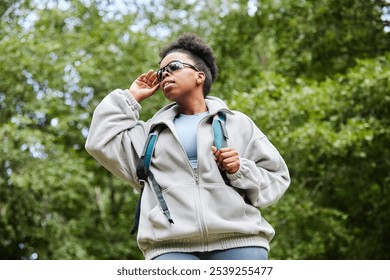 Vibrant low angle portrait of active African American woman as female tourist exploring forest with backpack and adjusting sunglasses against greenery copy space - Powered by Shutterstock