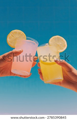 A vibrant, low-angle shot captures two hands holding up glasses of colorful lemonade, garnished with lemon slices, in a celebratory toast against a bright blue sky.