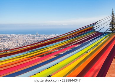 Vibrant multicolored hammocks at an elevated viewpoint overlooking the panoramic cityscape of Barcelona and the Mediterranean Sea. Bright travel image representing relaxation, summer holidays, tourism, and leisure in Catalonia, Spain.