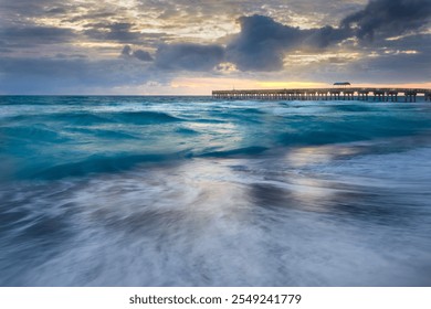 Vibrant ocean waves crash against the shore under a sunrise, with Lake Worth pier extending into the horizon. A blend of dynamic motion and serene colors captures the beauty of coastal mornings. - Powered by Shutterstock