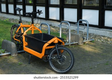 A vibrant orange cargo bike is secured at a bike rack beside a contemporary building. The bike features a spacious front cargo area, showcasing its utility for transporting goods. - Powered by Shutterstock