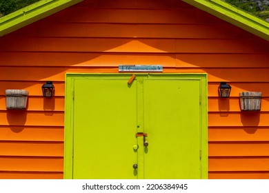 A vibrant orange and lime green wooden beach hut shelter. The exterior wall is a vibrant color with exterior lights and a sign: life is better near the water, over the main door of the building. 