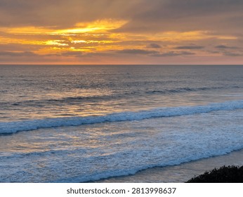 Vibrant orange sunset lighting up dramatic cloud layers above the Pacific Ocean with gentle ocean waves breaking along the Carlsbad California coast.