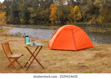 A vibrant orange tent set up by a serene riverbank, surrounded by autumn foliage. A wooden table with a thermos, cups, and apples is nearby, creating a cozy camping scene. - Powered by Shutterstock