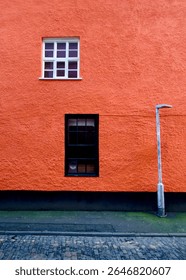 A vibrant orange wall features two windows, one white and one black, and a streetlight. The contrasting colors and textures of the wall, windows, and streetlight create a striking visual scene.