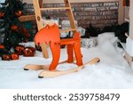 A vibrant orange wooden rocking horse stands in a cozy winter scene, surrounded by white snow and holiday decorations. Nearby, a ladder and Christmas ornaments enhance the festive atmosphere.