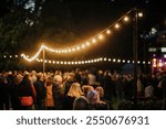 A vibrant outdoor evening gathering featuring a crowd under glowing string lights, set against a dark background with trees and a stage visible in the distance