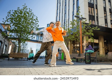 Vibrant Outdoor Scene Capturing Two Young Adults Joyfully Dancing In An Urban Setting, Showcasing Friendship And Modern Youth Culture Against A Background Of Contemporary Architecture And Greenery.