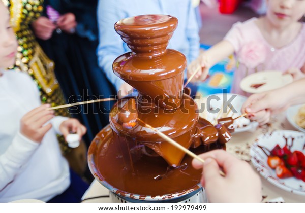 Vibrant Picture of Chocolate\
Fountain Fontain on childen kids birthday party with a kids \
playing around and marshmallows and fruits dip dipping into\
fountain