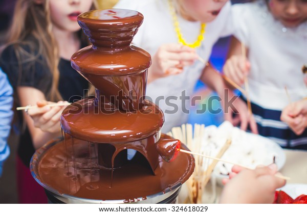 Vibrant Picture of Chocolate\
Fountain Fontain on childen kids birthday party with a kids playing\
around and marshmallows and fruits dip dipping into fountain\
