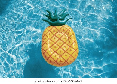 A vibrant pineapple-shaped inflatable pool float rests on the surface of a clear, blue swimming pool, with sunlight creating shimmering patterns on the water.