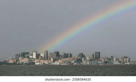 A vibrant rainbow arches over a sprawling urban skyline of Rio de Janeiro, Brazil, with high-rise buildings and modern architecture reflected in the calm waters of Guanabara Bay - Powered by Shutterstock