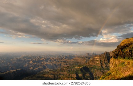 A vibrant rainbow cuts through dramatic storm clouds over the vast canyons and jagged peaks of Simien Mountains National Park during golden hour in Ethiopia.