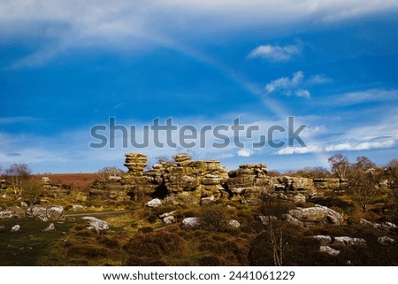 Vibrant rainbow over a rocky landscape with scattered boulders and lush greenery under a blue sky with clouds at Brimham Rocks, in North Yorkshire