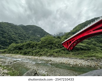 A vibrant red bridge gracefully arches over a lush green valley with a serene river running below, set against a backdrop of misty mountains and dramatic clouds. - Powered by Shutterstock