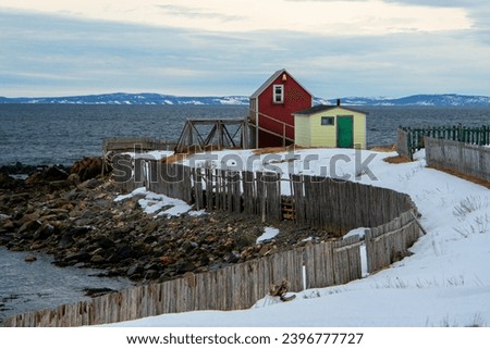 A vibrant red colored barn with a window in the loft and a yellow shed with a green wooden door on a point overlooking the ocean. A seawall of wood logs lines the coastline covered in white snow.