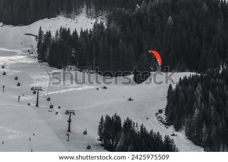 A vibrant red paraglider soars above a snowy mountain landscape, embodying the concept of freedom and adventure. Ski lifts and skiers dot the pristine slopes, contrast of tranquility and exhilaration