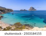 Vibrant scene of Cala Hort beach, Ibiza, featuring anchored boats and the stunning contrast of emerald waters against the rugged cliffs. Es Vedra and Es Vedranell islands enhance the coastal view