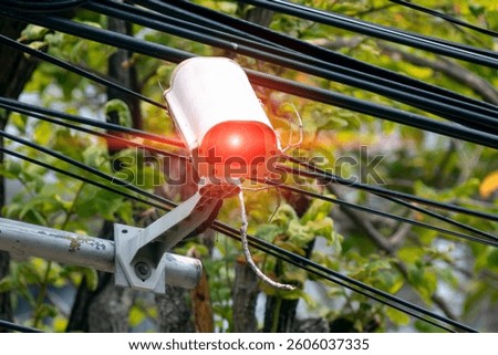 A vibrant security camera with a glowing red light mounted on power lines, surrounded by lush green foliage, showcasing modern surveillance technology in urban settings.