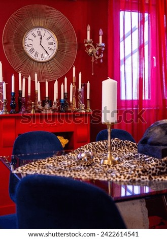 In a vibrant setting, a red room hosts a leopard-print table and plush blue chairs, highlighted by a golden sunburst clock and an array of eclectic candlesticks.


