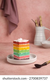 A vibrant slice of rainbow layer cake with creamy white frosting, served on a small gray plate. The dessert stands out against the soft, dusty pink background with bamboo handled utensil