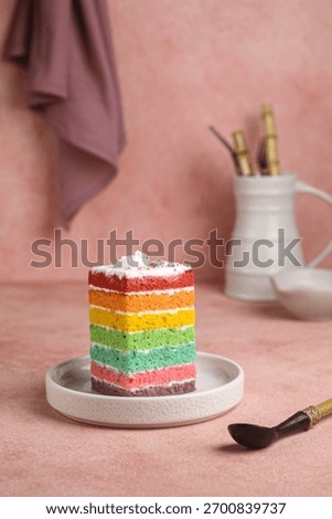 A vibrant slice of rainbow layer cake with creamy white frosting, served on a small gray plate. The dessert stands out against the soft, dusty pink background with bamboo handled utensil
