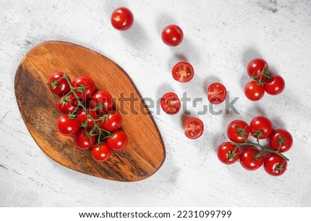 Vibrant small red tomatoes with green vines on wooden chopping board, white stone table under, view from above