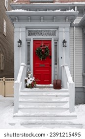 A vibrant solid red door with side glass windows and a Christmas wreath hanging on it. The entrance to the white wooden traditional house has steps, a decorative pot of flowers, and a snow shovel.