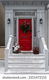 A vibrant solid red door with side glass windows and a Christmas wreath hanging on it. The entrance to the white wooden traditional house has steps, and a decorative pot of flowers and a snow shovel. 