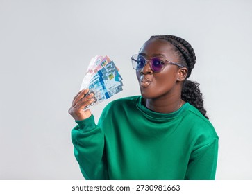 A vibrant studio portrait features a young black African woman with a confident, playful expression, holding a fan of Ghanaian Cedi banknotes. She wears a rich forest-green sweatshirt and stylish purple-tinted glasses, with her hair neatly styled in braids transitioning into a ponytail. Set against a clean, neutral grey background, the lighting is soft and professional, highlighting the texture of the currency and her features. The image exudes a sense of financial success, youth, and modern African style