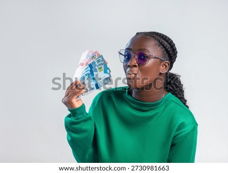 A vibrant studio portrait features a young black African woman with a confident, playful expression, holding a fan of Ghanaian Cedi banknotes. She wears a rich forest-green sweatshirt and stylish purple-tinted glasses, with her hair neatly styled in braids transitioning into a ponytail. Set against a clean, neutral grey background, the lighting is soft and professional, highlighting the texture of the currency and her features. The image exudes a sense of financial success, youth, and modern African style