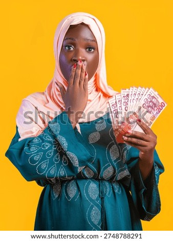 A vibrant studio portrait features a young Black woman against a bright yellow background. She wears a light peach hijab and a teal patterned kaftan. The ladies expression is one of theatrical shock, with her right hand covering her mouth and her eyes wide. In the left hand, she fans out several 200 Ghana Cedi banknotes, showcasing the red and purple currency. The high-contrast lighting emphasizes her surprise and the wealth she is holding, creating a bold, energetic visual.