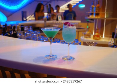 A vibrant and stylish bar setting featuring two colorful cocktails. The drinks, beautifully presented on the bar counter, are illuminated by striking blue neon lights in the background. - Powered by Shutterstock