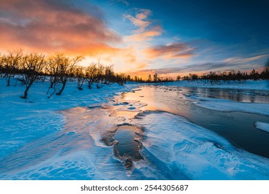 The vibrant sunrise casts a warm glow over a snowy river landscape, where silhouetted trees frame the scene. The water reflects orange and blue hues of the sky. - Powered by Shutterstock